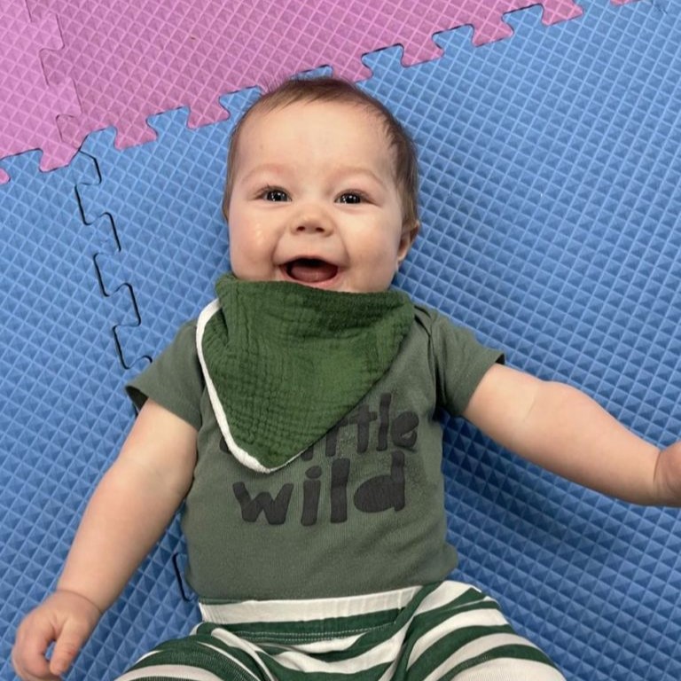 A small child lies on a textured foam play mat with blue and purple interlocking tiles. The child is wearing a green outfit with a matching green bib and striped green-and-white pants, along with a shirt that reads “little wild.” One arm is extended outward while resting on the mat.