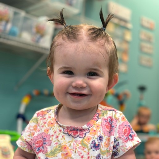 Young child with two small ponytails wearing a floral shirt, sitting in a colorful playroom with toys and shelves in the background.
