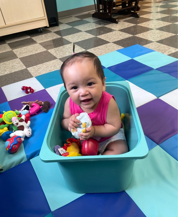 Infant sitting in a sensory bin surrounded by colorful toys in the Club Baby program at The Clubhouse childcare center.