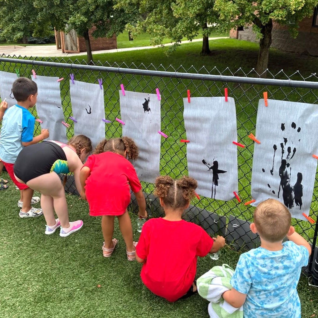 Children creating outdoor art at The Club House summer program. Children enjoying a hands-on outdoor art activity at The Club House, using water and paint to create artwork on large paper panels attached to a fence during a summer program.